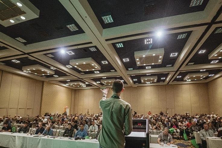 A person speaking on a stage in a conference room.