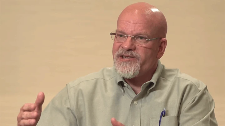 A bald man with a white beard and glasses wearing a tan button-up shirt gestures with both hands as he talks.