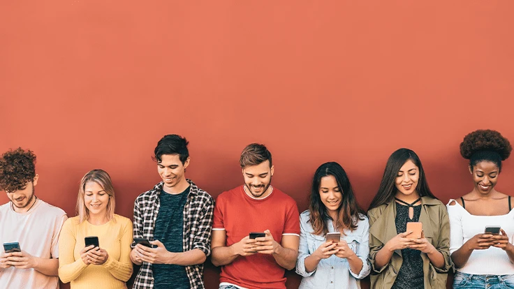 Seven people on their phones lean on an orange wall.