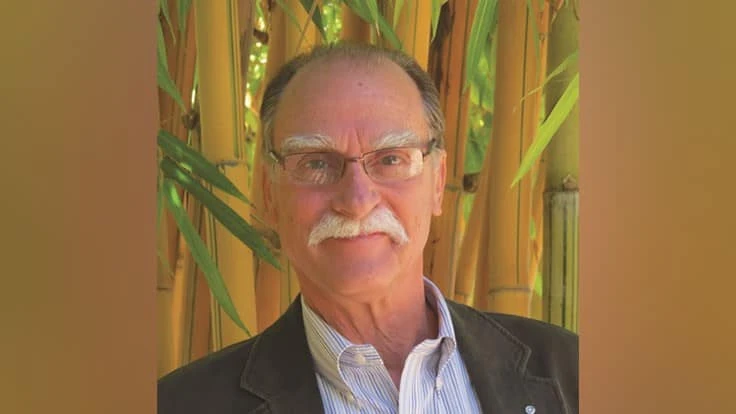 A man in glasses and gray hair smiles at the camera in front of a wall of bamboo