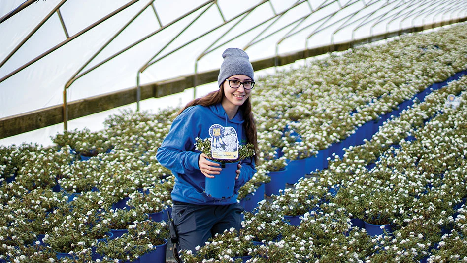 A Woman With Glasses In A Blue Sweatshirt and Gray Hat Surrounded By Potted Plants