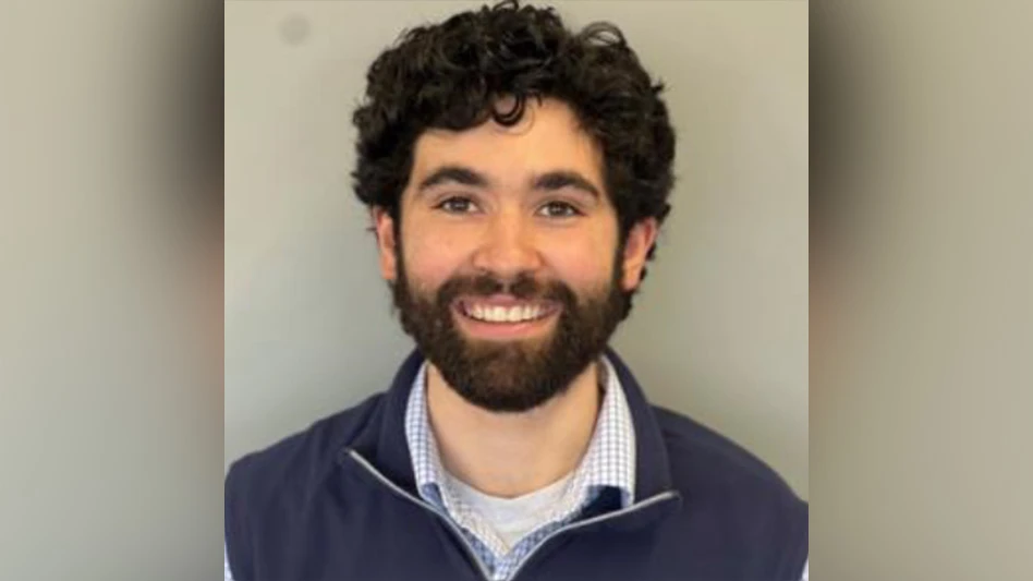 A white man with black hair and a black beard wearing a navy blue vest over a blue and white checked button-up shirt and a white T-shirt smiles in a headshot photo, from the shoulders up.