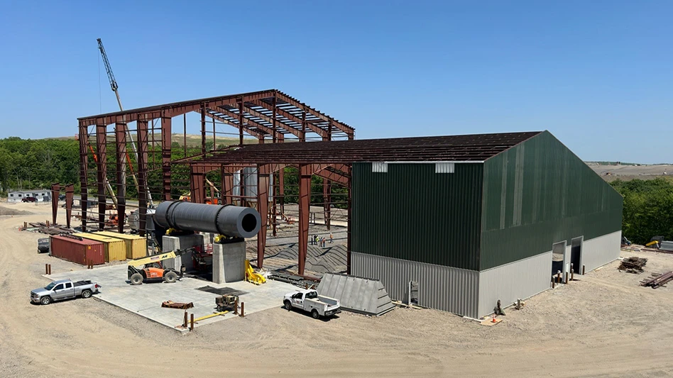 A large building is under construction and partially built. The front is a dark green and gray barn-looking structure, while the back is only reddish-brown framing. There are a few trucks and construction equipment throughout the photo, which includes a dirt ground, green trees and a blue sky.