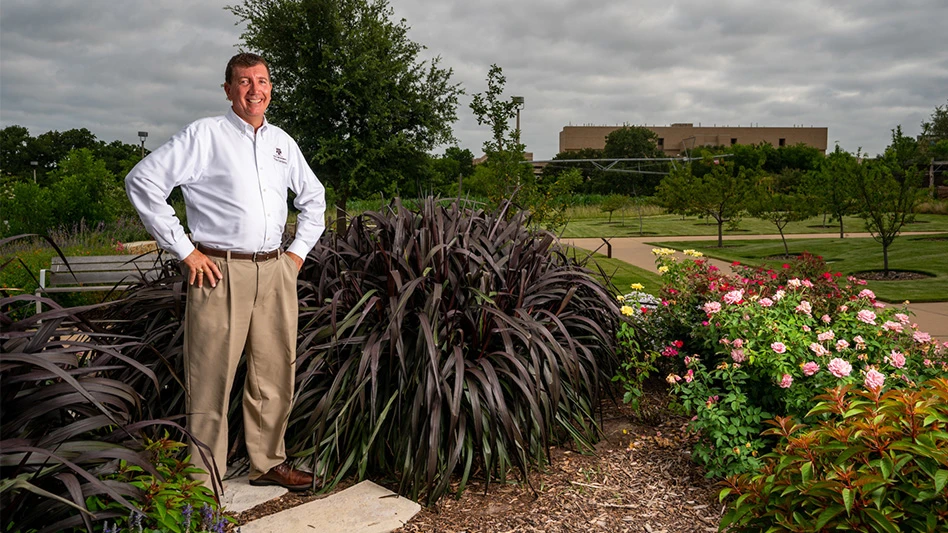 A smiling man with dark hair wearing a white long-sleeve dress shirt and khaki pants poses with his hands on his hips. He stands in a garden on a university campus.