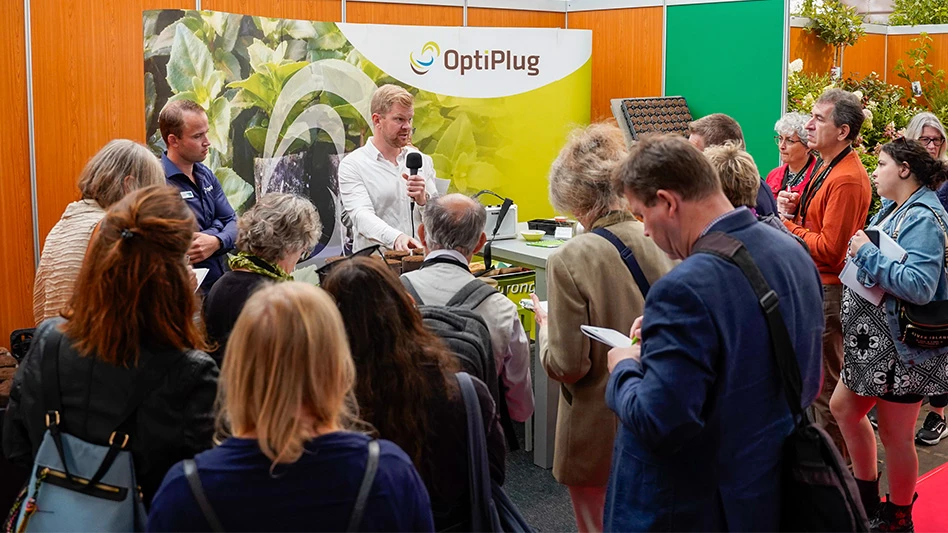 A man speaks to a small crowd of people in front of a yellow, orange and green OptiPlug display.
