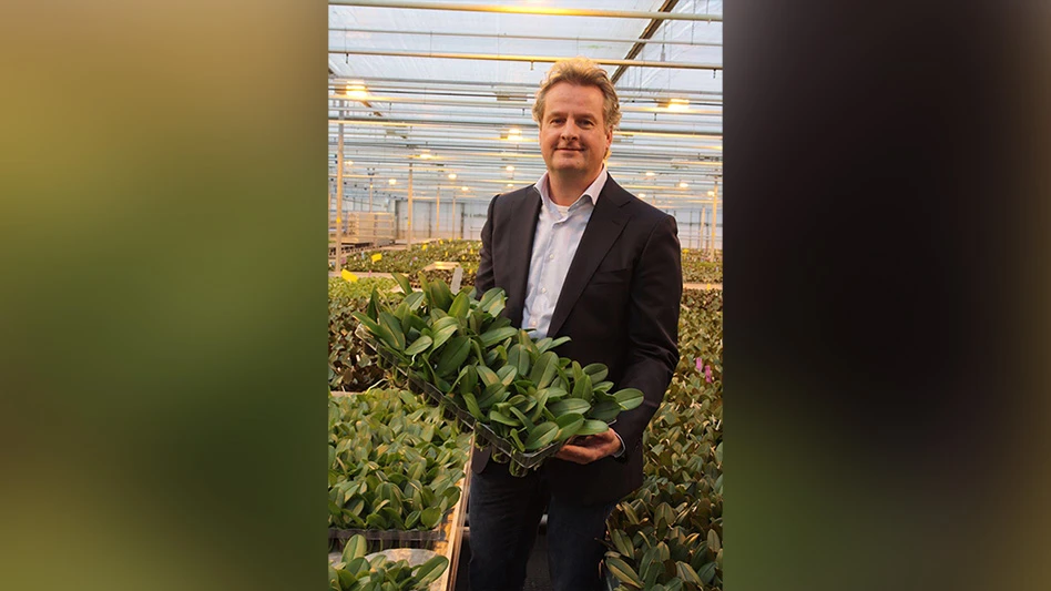 A man wearing a black suit jacket and light blue shirt holds a tray of plants with green leaves in a greenhouse, with more of the same plants behind him/