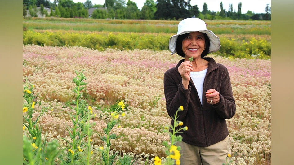 A smiling woman with short black hair wearing a white hat, white shirt, brown jacket and khaki pants holds a green seedling standing in a field of pink, tan and green flowers and plants.