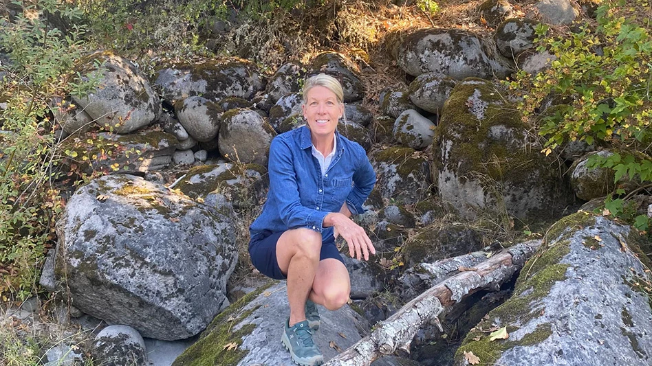 A smiling woman with blonde hair wearing a blue denim long-sleeve button-up shirt, navy blue shorts and gray hiking shoes rests her right arm on her right knee as she kneels amid several large gray mossy rocks on a hill.