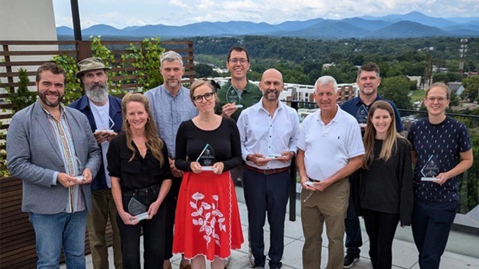 A group of 11 people smile and hold their awards as they pose for a photo on a patio, with green trees and mountains visible in the background.