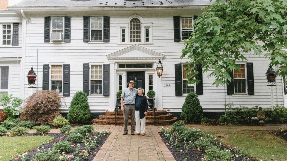 A man and woman with their arms around each other stand in front of a white house with black shutters and a brick walkway and steps, with green plants along the path and in front of the house.