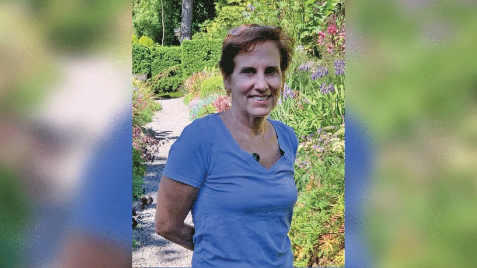 A smiling woman with short brown hair wearing a periwinkle-colored T-shirt stands with her arms behind her back and poses in front of a garden.