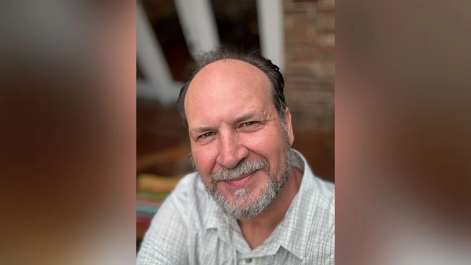 A smiling man with short dark hair and a short gray beard wearing a tan and white plaid shirt takes a selfie from a high angle.