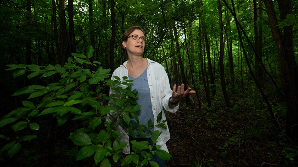A woman with short brown hair wearing a blue t-shirt, white button-up jacket and glasses gestures with her left hand as she talks. She stands surrounded by trees in a nature preserve.