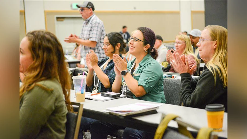 People in a room sitting at tables in a conference room smile and applaud.