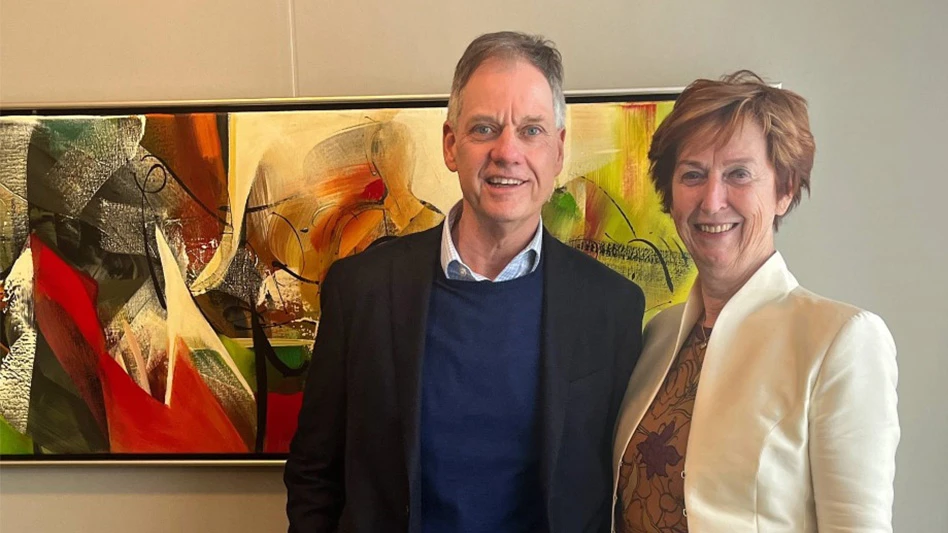 A photo of two smiling people posing in front of a piece of art. The man on the left has short dark gray hair and wears a blue shirt, navy blue sweater and black suit jacket. The woman on the right has short red hair and wears a brown floral top and cream jacket.