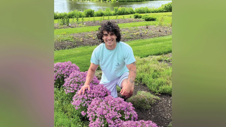 A man with long brown hair kneeling behind a row of pink shrubs.