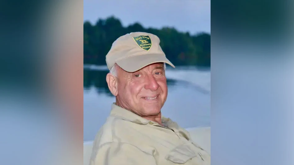 A smiling man wearing a tan baseball cap and tan shirt and sitting beside a pond.