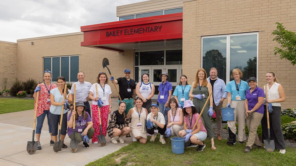A group of people stand in front of a high school with shovels, plant containers and soil.