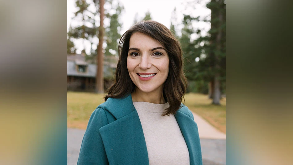 A smiling woman with shoulder-length brown hair wearing a cream sweater and teal jacket. Trees and a building are visible in the background.