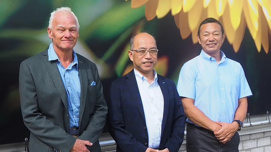 Three smiling men in front of a green wall with a yellow flower.