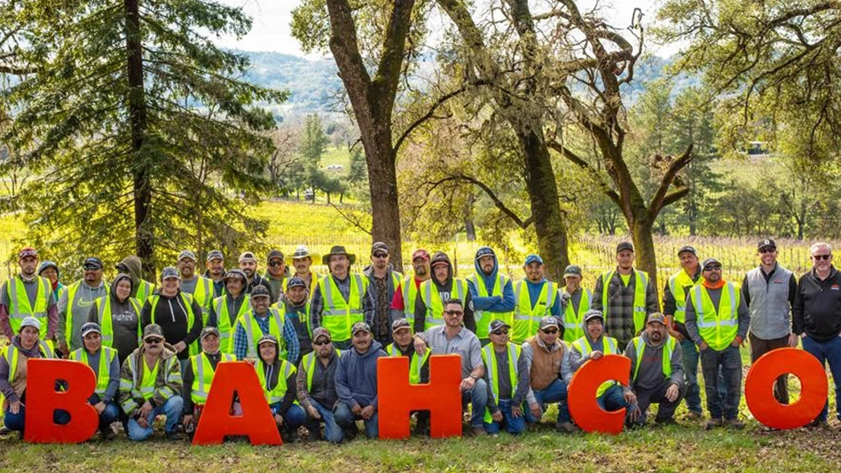 A crowd of people, many wearing neon yellow safety vests, pose in front of red letters reading BAHCO. They stand in front of green trees and fields.