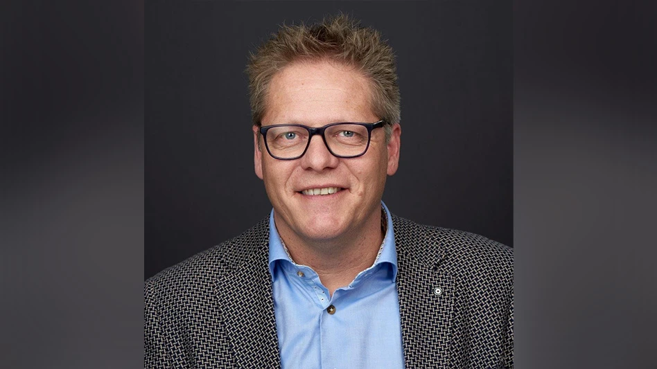 A smiling man with short spiky light brown hair wearing black glasses, a light blue button-up shirt and a black and white checked jacket.