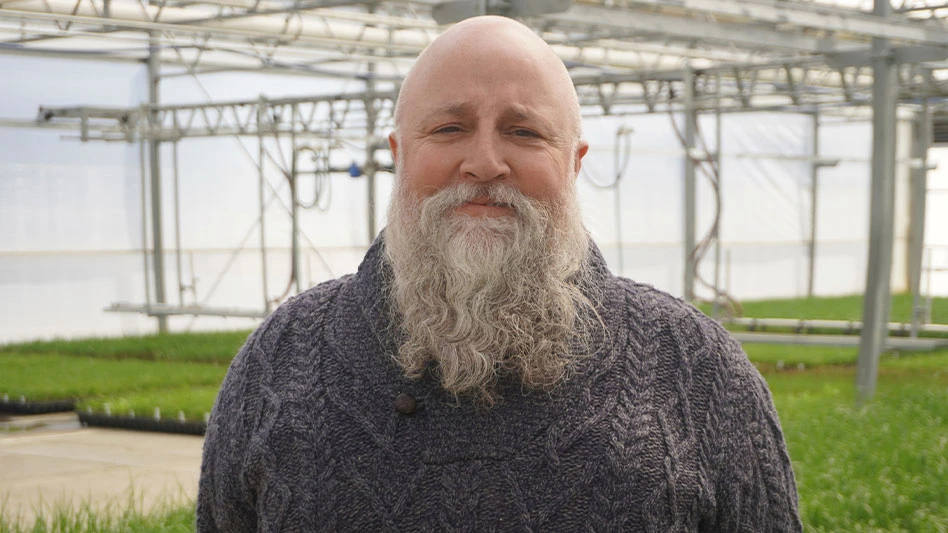 A bald man with a long gray beard who's wearing a gray sweater and standing inside a greenhouse.