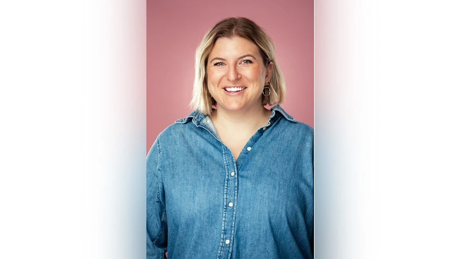 A smiling woman with short blonde hair in a blue button-down shirt in front of a pink background.