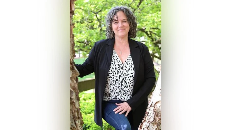 A smiling woman with short dark gray hair and wearing a white top with black spots, a black jacket and blue jeans poses next to a tree.