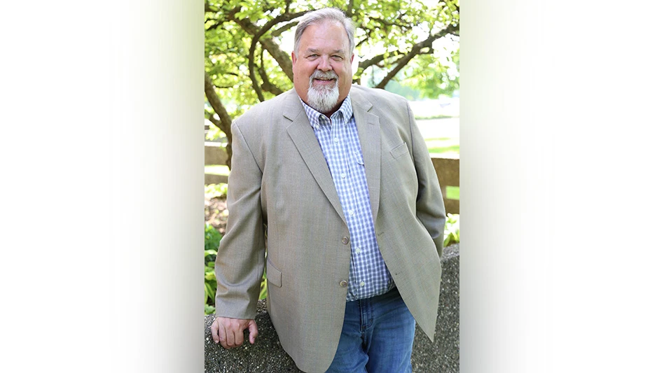 A smiling man with short white hair and a short white beard wearing a gray suit jacket, blue and white plaid button-up shirt and blue jeans leans against a gray stone wall in front of a tree.