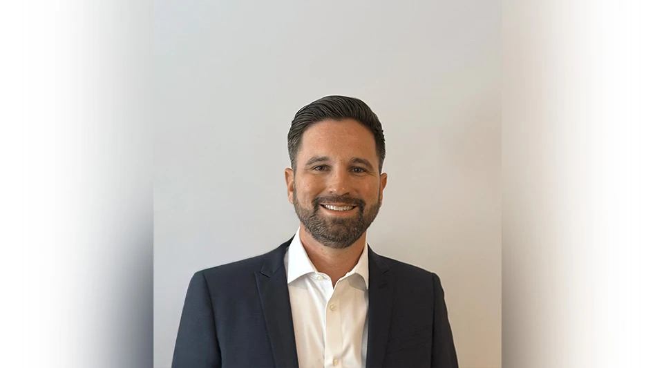 A smiling man in a white dress shirt and black suit jacket standing in front of a white wall.
