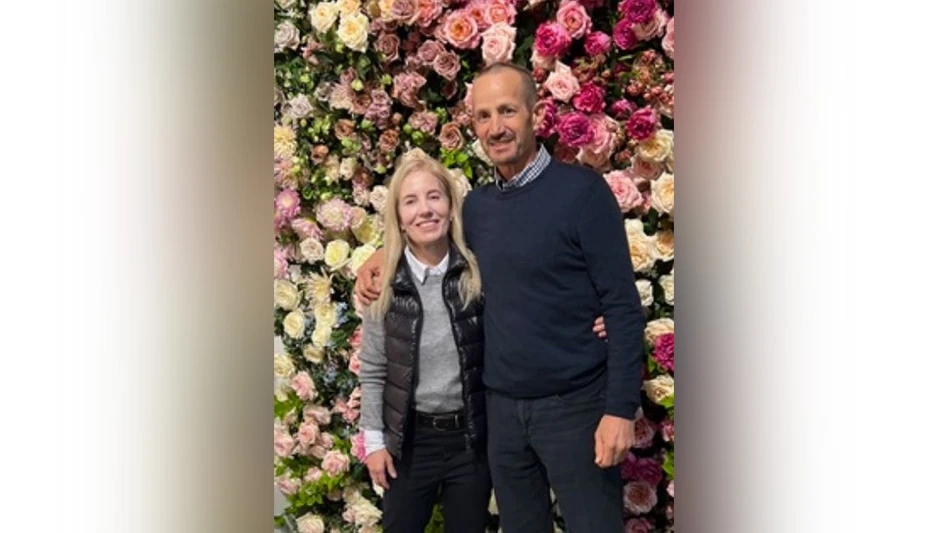 A smiling man and woman pose in front of a background made up of pink and white flowers.