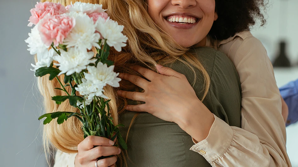 A person visible from the nose up holding a pink and white bouquet of flowers and wearing a tan long-sleeved shirt hugs a person wearing an olive green shirt with long blonde hair who's facing away from the camera.