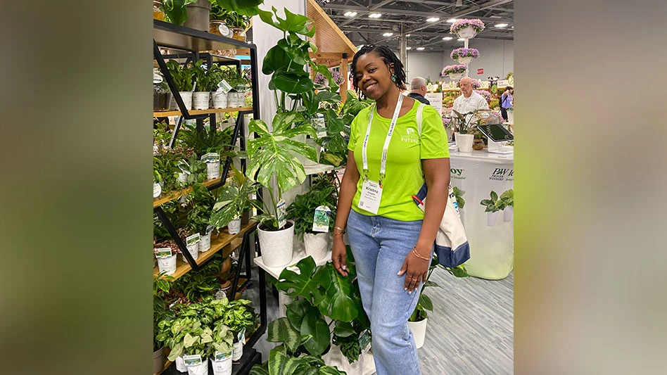 A student wearing a green shirt and blue jeans stands beside a display of houseplants at a horticulture industry trade show.