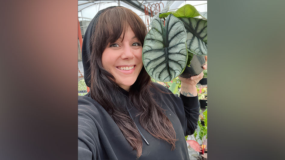 A smiling woman with long brown hair and wearing a black hoodie with the hood up holds up a giant green leaf.