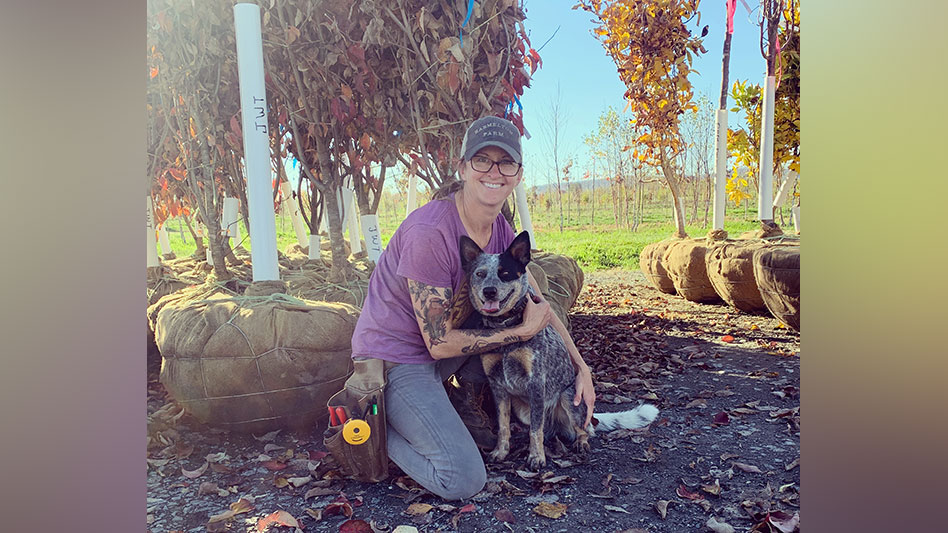 A smiling woman with a dog kneels in front of trees.