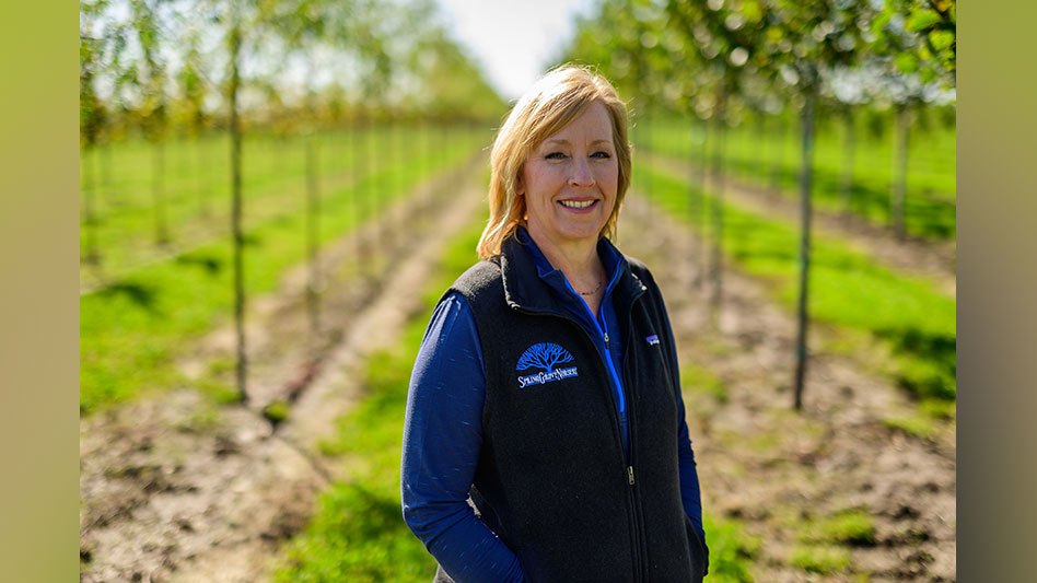 A smiling woman in a blue shirt and vest walking through rows of trees at a nursery.