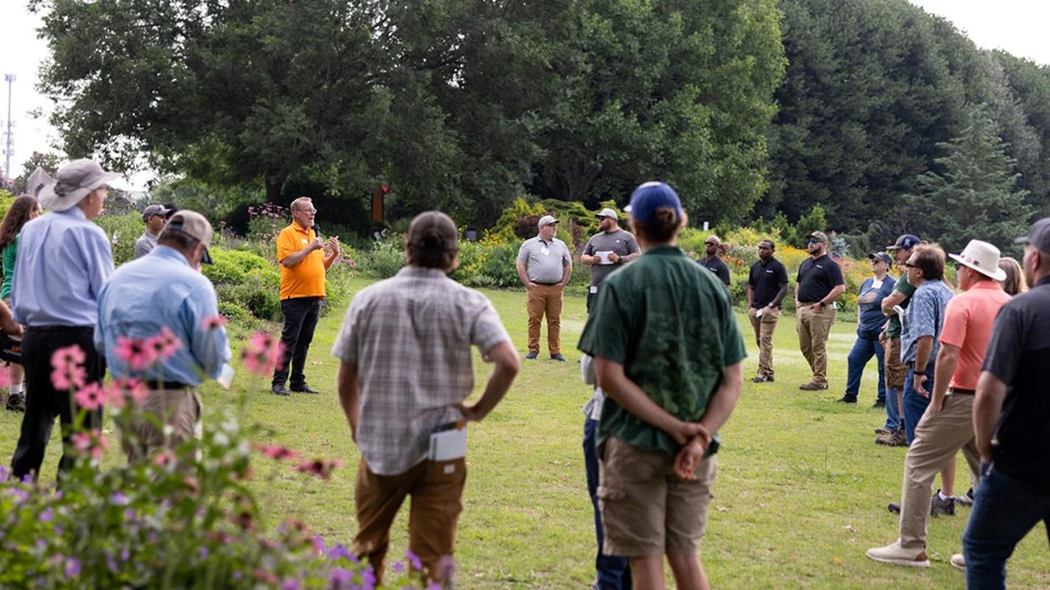 A group of people listen to a man holding a microphone talking while they stand in a field.