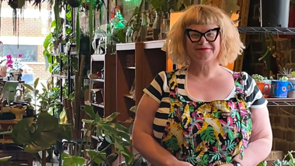 A smiling woman with chin-length blonde hair and wearing black glasses, a black and white striped shirt and a black apron with green leaves on it stands in a shop full of plants on wooden shelves.