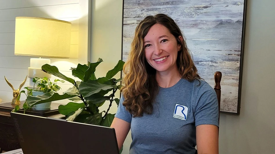 a woman in a gray t shirt smiles at the camera in front of a laptop