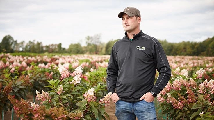 A man in blue jeans, a black quarter-zip, and a cap stands in a field of light pink flowers
