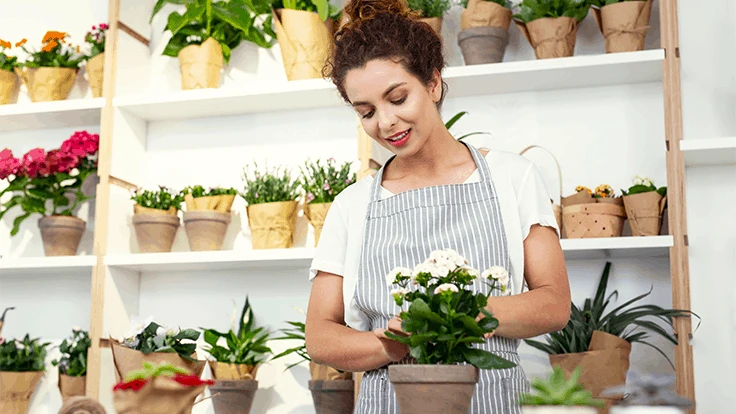a woman in a white t shirt and striped apron stands in front of shelves of potted plants